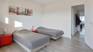 A bedroom with two beds, a red nightstand, a lamp, and a cityscape painting on the wall. - Appartement Panorama - Welkeys (Villeneuve-Loubet)