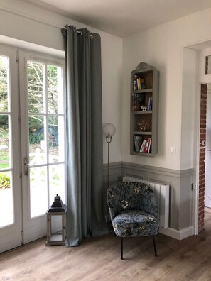 Quarto com uma janela grande, cortina cinza, estante de livros, poltrona estampada e um pequeno candeeiro.