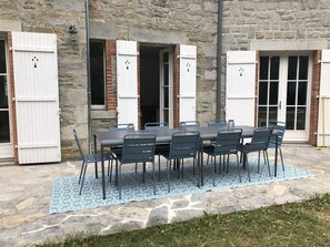 Um edifício de pedra com uma mesa e cadeiras no pátio, janelas com persianas brancas e chão de pedra.
