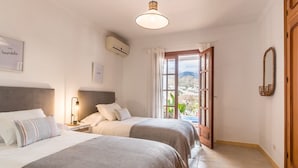 A bedroom with two beds, a bedside table with a lamp, an air conditioning unit, and a window with a view of a mountainous landscape.