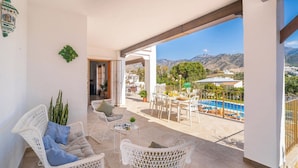 A balcony with white wicker furniture, a small table, and a view of a swimming pool and mountains.