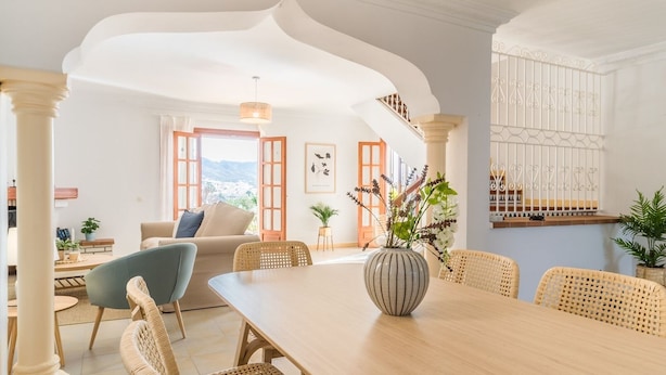 A spacious living area with a wooden dining table, wicker chairs, a sofa, and a large window with a view.