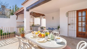 An outdoor dining area with a table set for breakfast, surrounded by white chairs, a wooden pergola, and a white wall with a door.