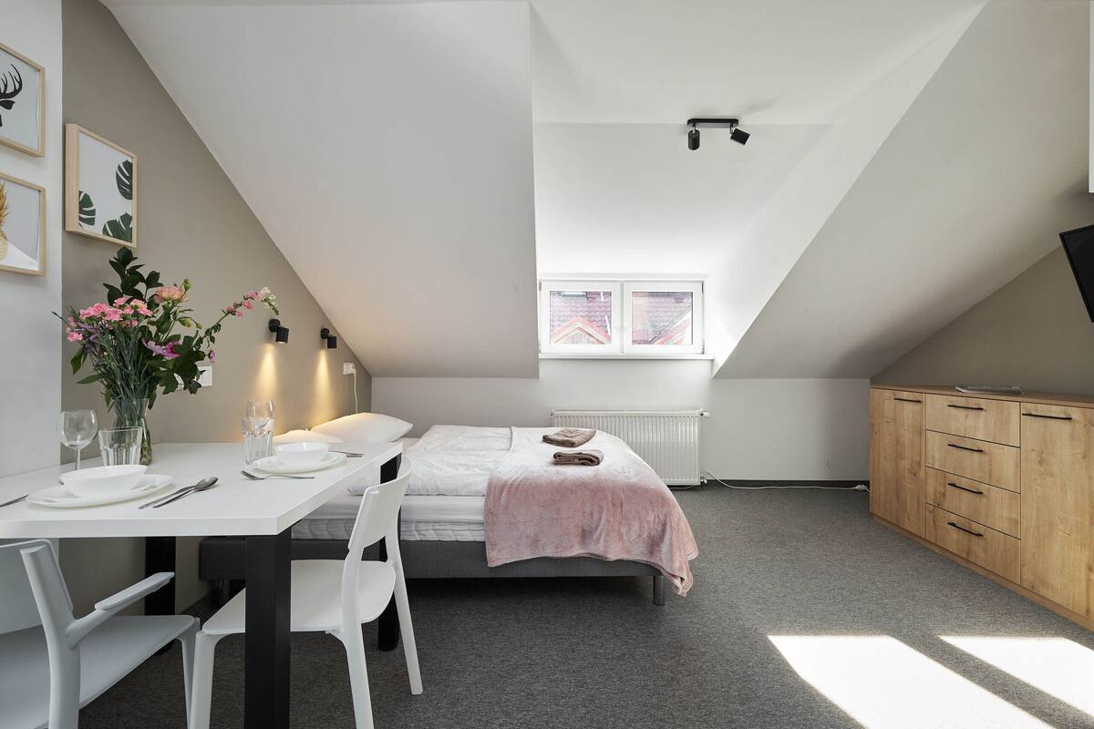 Un dormitorio moderno con techo inclinado, una cama con manta rosa, un armario de madera, un ventanal pequeño y una zona de comedor con mesa preparada para dos.