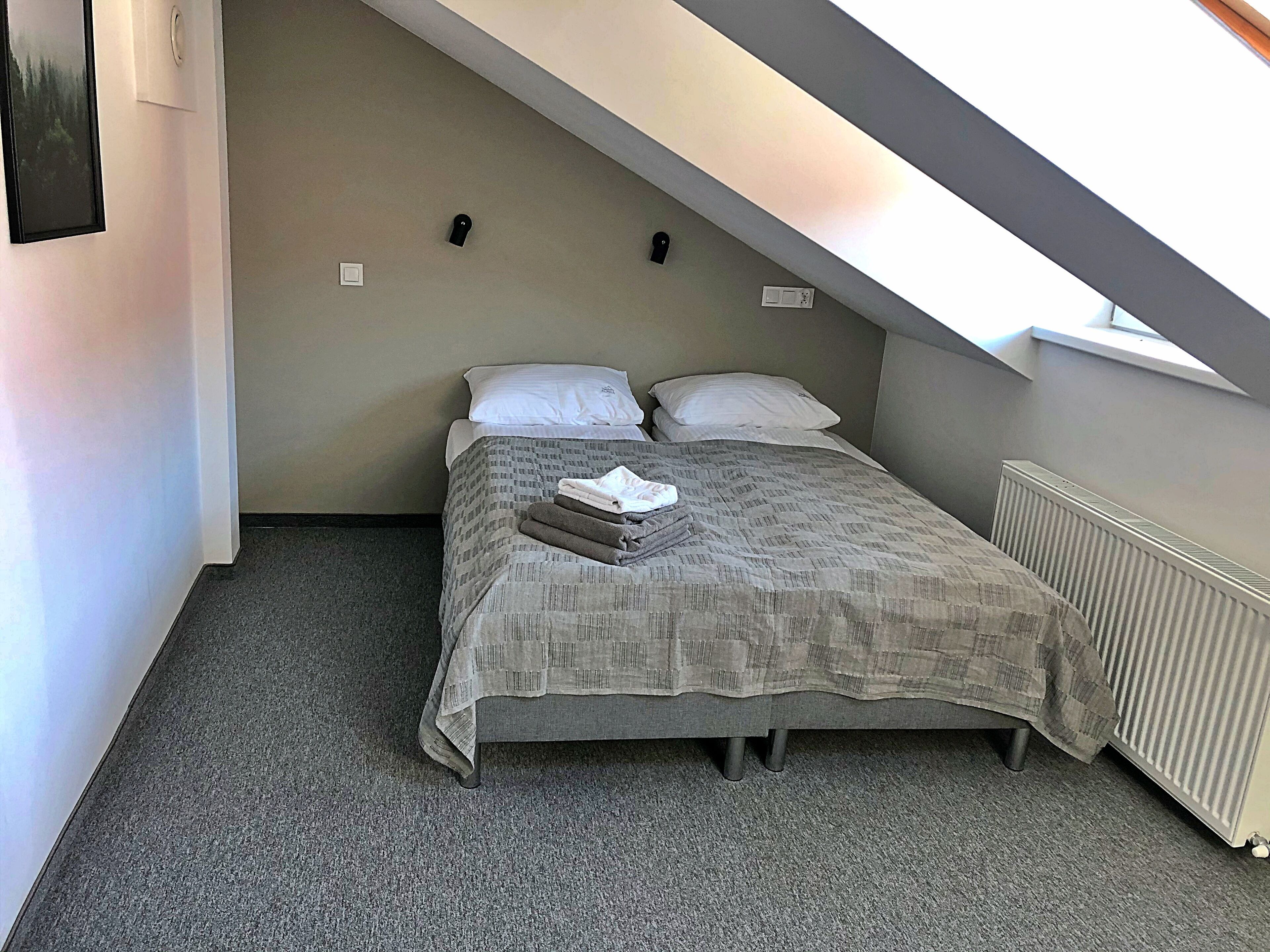 Un dormitorio pequeño y acogedor con techo inclinado, una cama con colcha gris estampada, dos almohadas y toallas dobladas con cuidado sobre la cama.