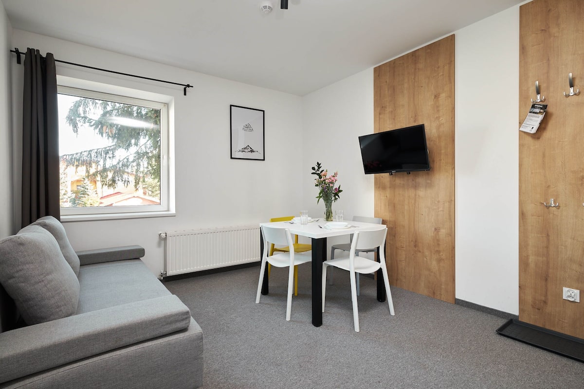 Una habitación moderna con un sofá gris, una mesa blanca con sillas, un armario de madera, un televisor montado y una ventana con cortinas.