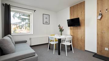 A modern room with a grey sofa, a white table with chairs, a wooden wardrobe, a mounted TV, and a window with curtains.