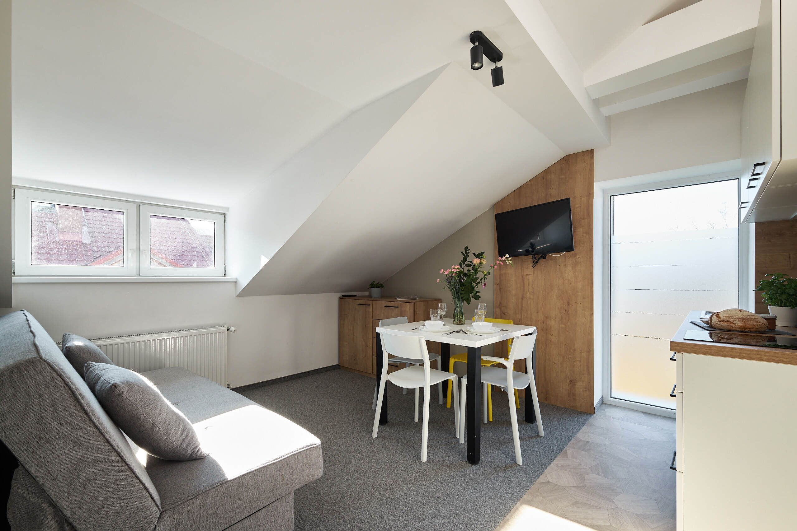 Un dormitori tipus loft modern amb sostre inclinat, televisió integrada, zona de menjador amb taula i cadires, i un sofà amb coixins.