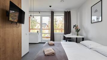 A hotel room with a bed, a desk, a chair, a TV mounted on the wall, and a window with curtains.