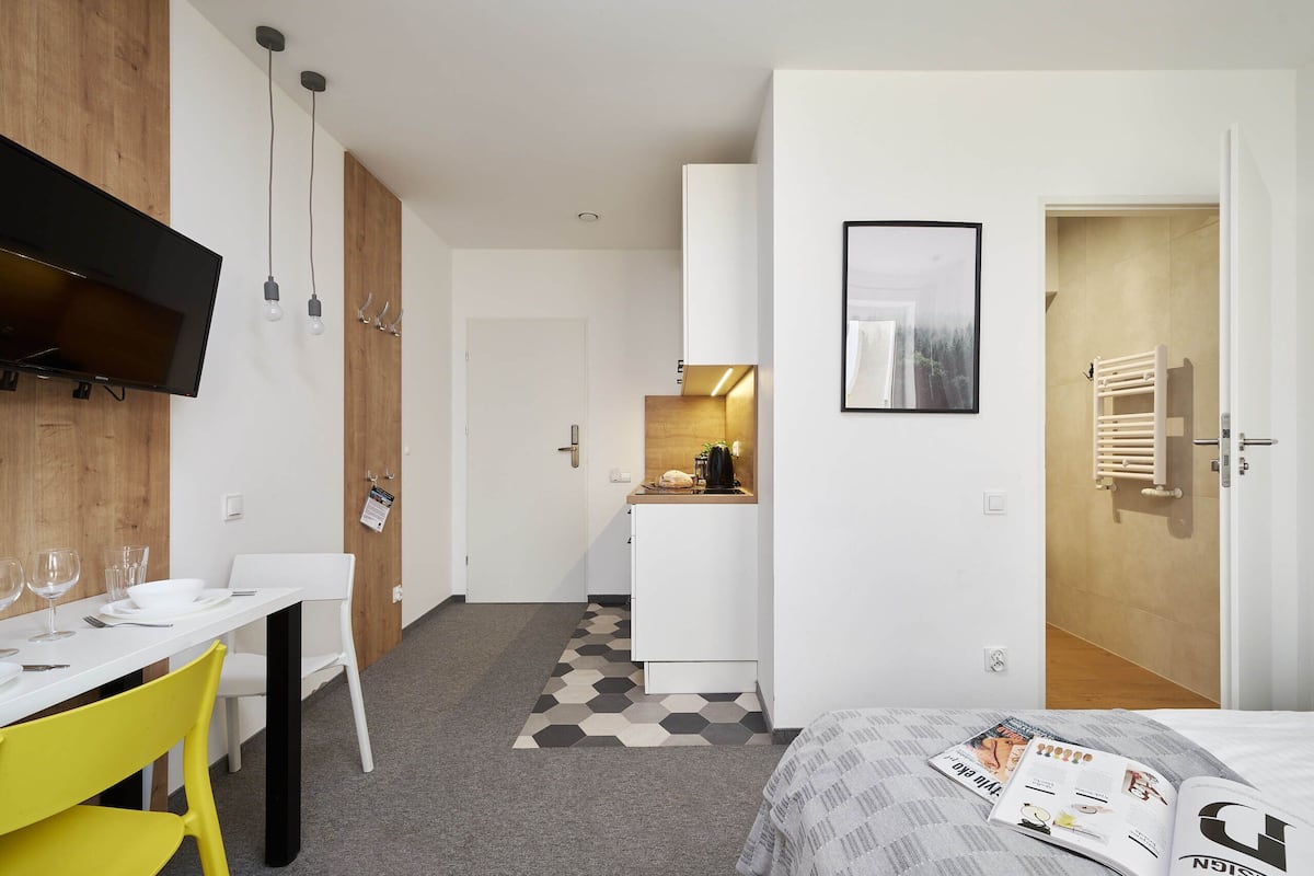 Una habitación de hotel moderna con una cama, una zona de comedor con una mesa y sillas, una kitchenette y un televisor montado en la pared.