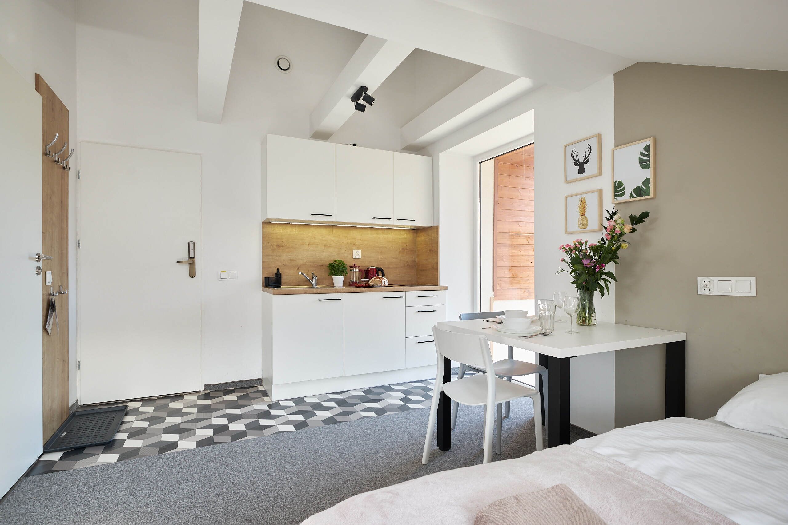 A modern hotel room with a bed, a dining table with chairs, a small kitchen area, and a bathroom.