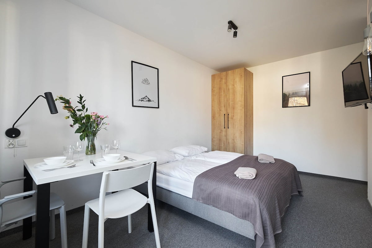 Una habitación de hotel moderna con una cama, un escritorio con una silla, una mesita con un jarrón de flores y dos cuadros en la pared.