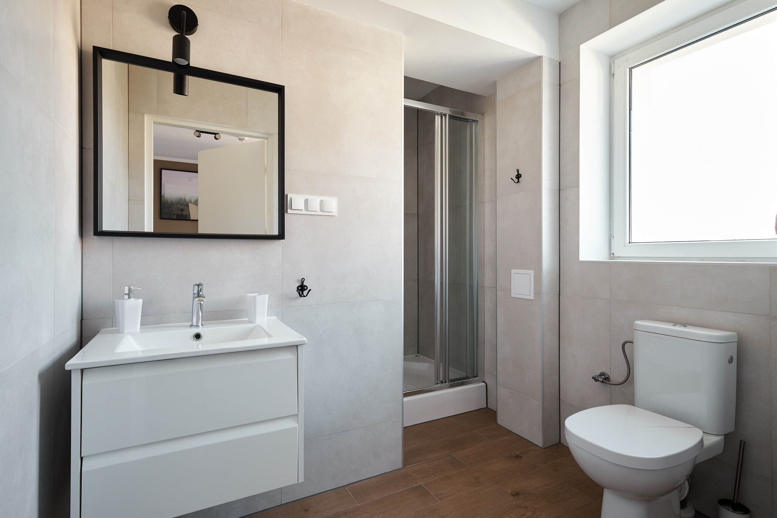 A modern bathroom with a white sink, mirror, and toilet, a shower area, and a window.