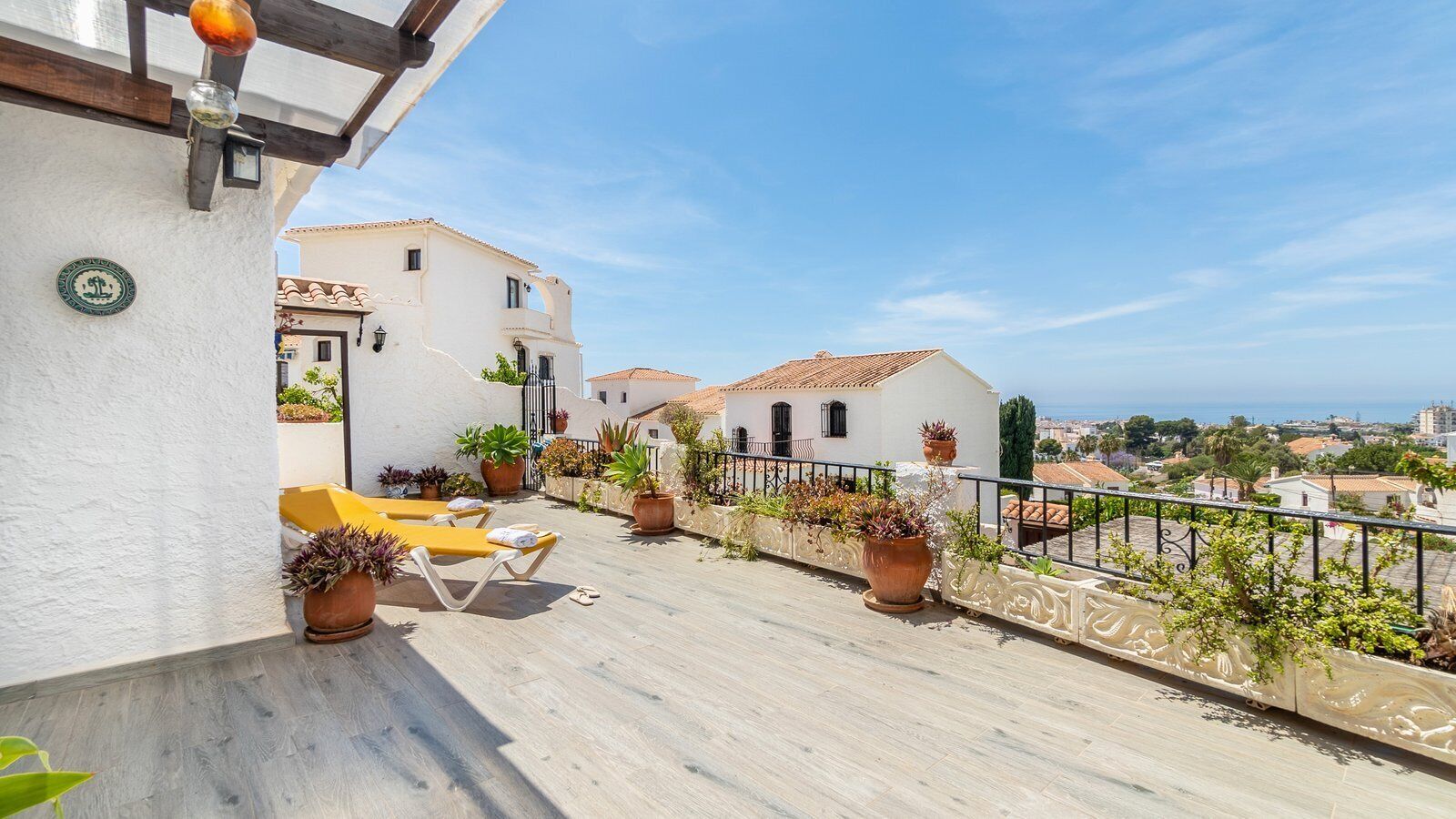 A rooftop terrace with sun loungers, potted plants, and a view of the sea and surrounding buildings.