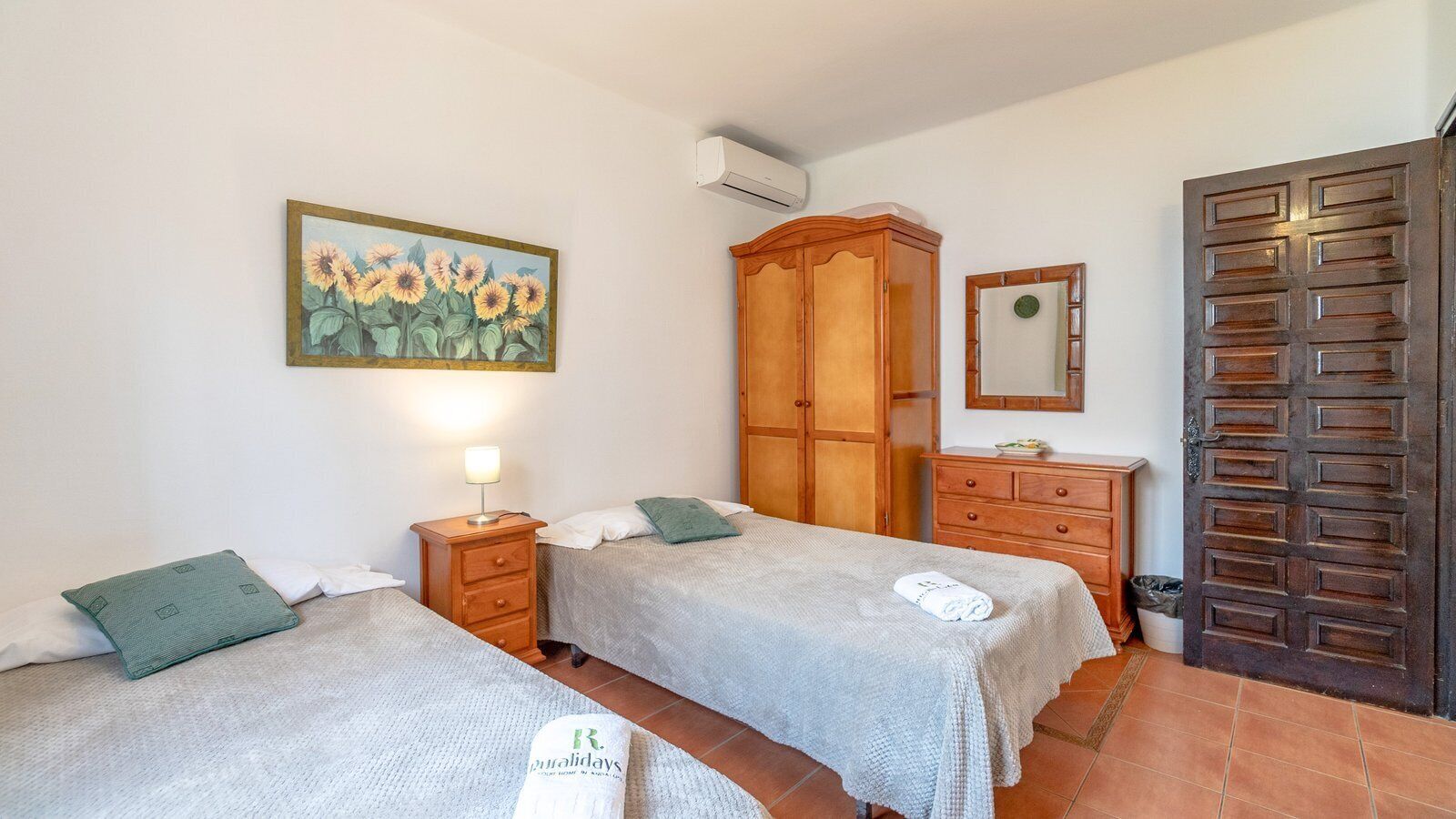 Een slaapkamer met twee bedden, een houten garderobe, een nachtkastje met een lamp, een spiegel en een deur.