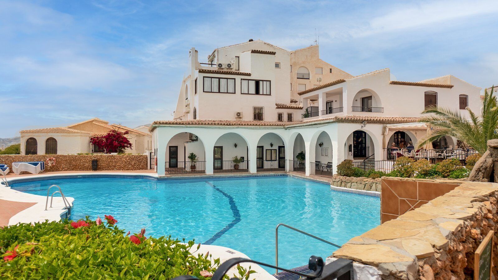 Un'area piscina con una grande vasca, un portico coperto con archi e un edificio con numerosi balconi.