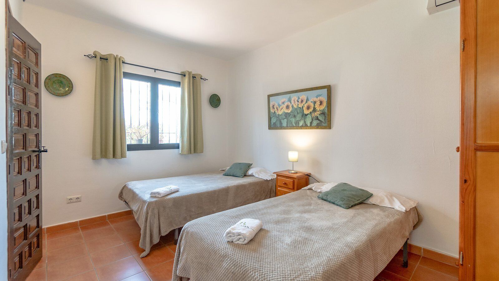 A room with two beds, a window with curtains, a painting of sunflowers, and a wooden door.