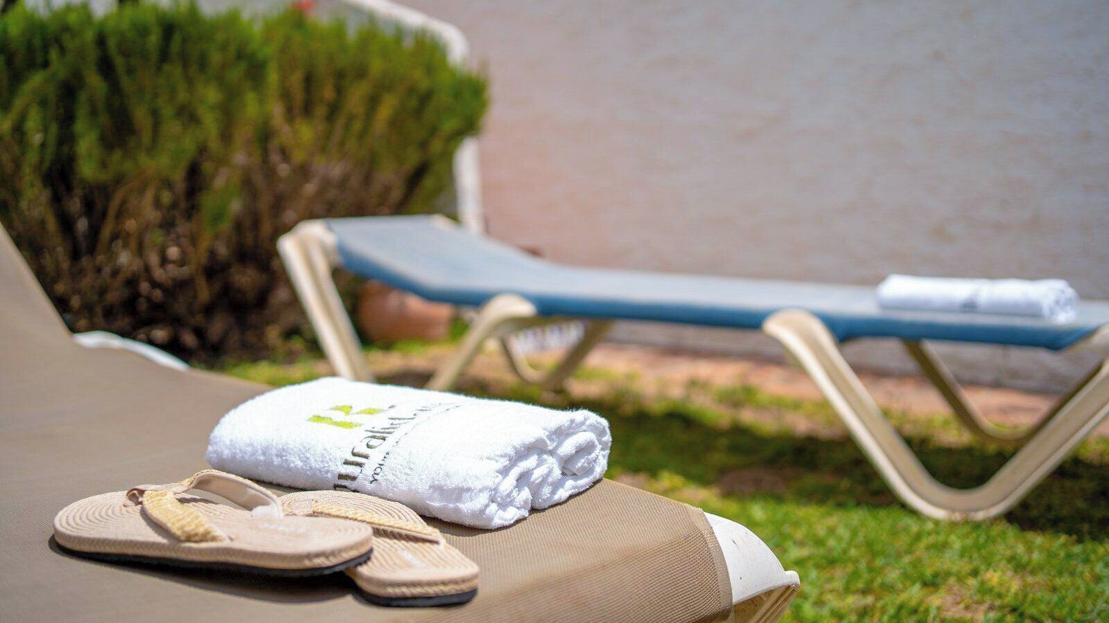 A sun lounger with a towel and flip-flops, a small table with a towel, and a neatly trimmed hedge in the background.