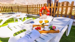 Ein sonnendurchfluteter Außenbereich mit weißen Liegestühlen, einem Holztisch mit Frühstücksgebäck und einem Holzzaun im Hintergrund.