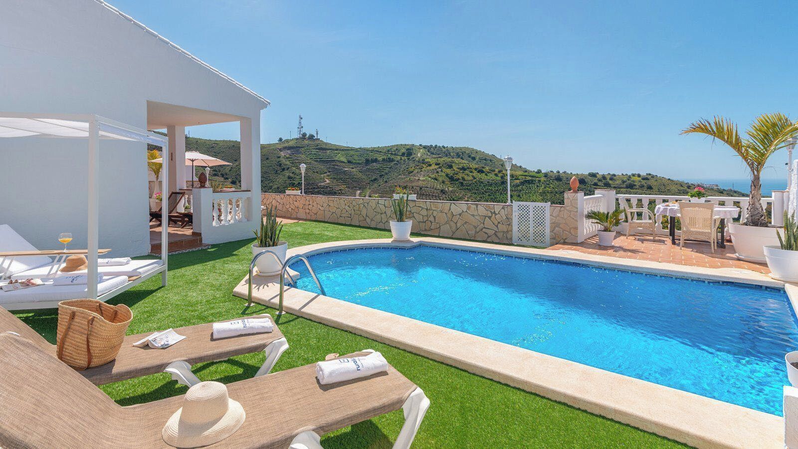 A pool area with lounge chairs, a covered seating area, and a view of the hills.