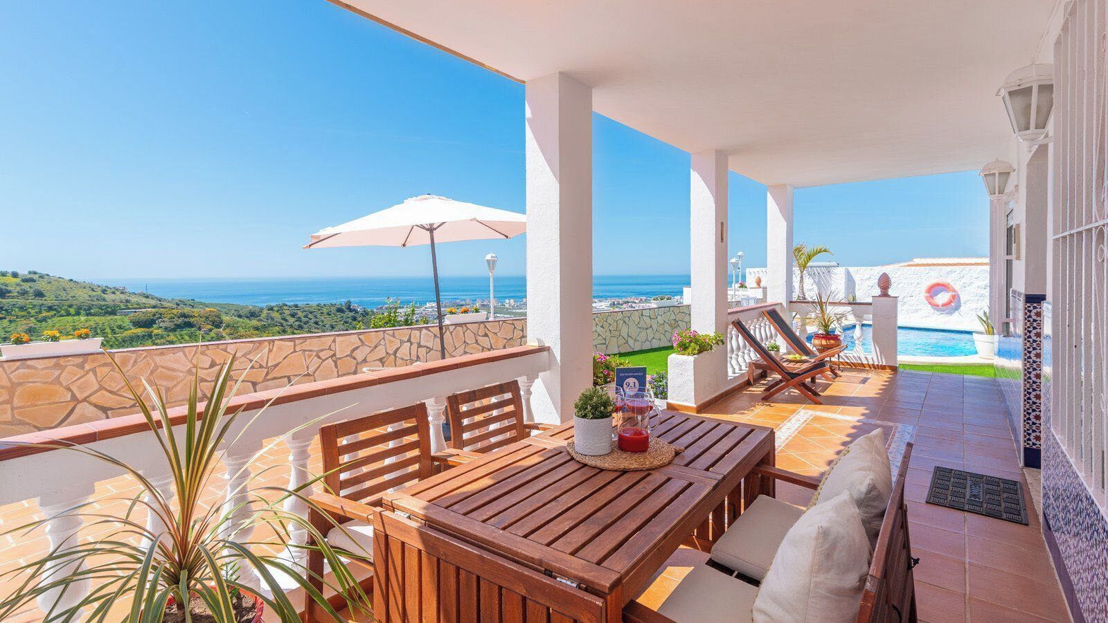 Een houten tafel en stoelen op een balkon met een stenen muur, uitzicht op de zee en een zwembad.