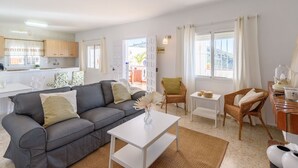 Una sala de estar moderna con un sofá gris, sillas de mimbre, una mesa de centro blanca y una cocina al fondo.