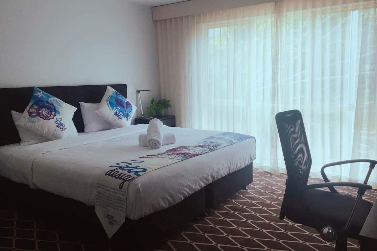 A hotel room with a large bed, a chair, a nightstand, and a window with sheer curtains.