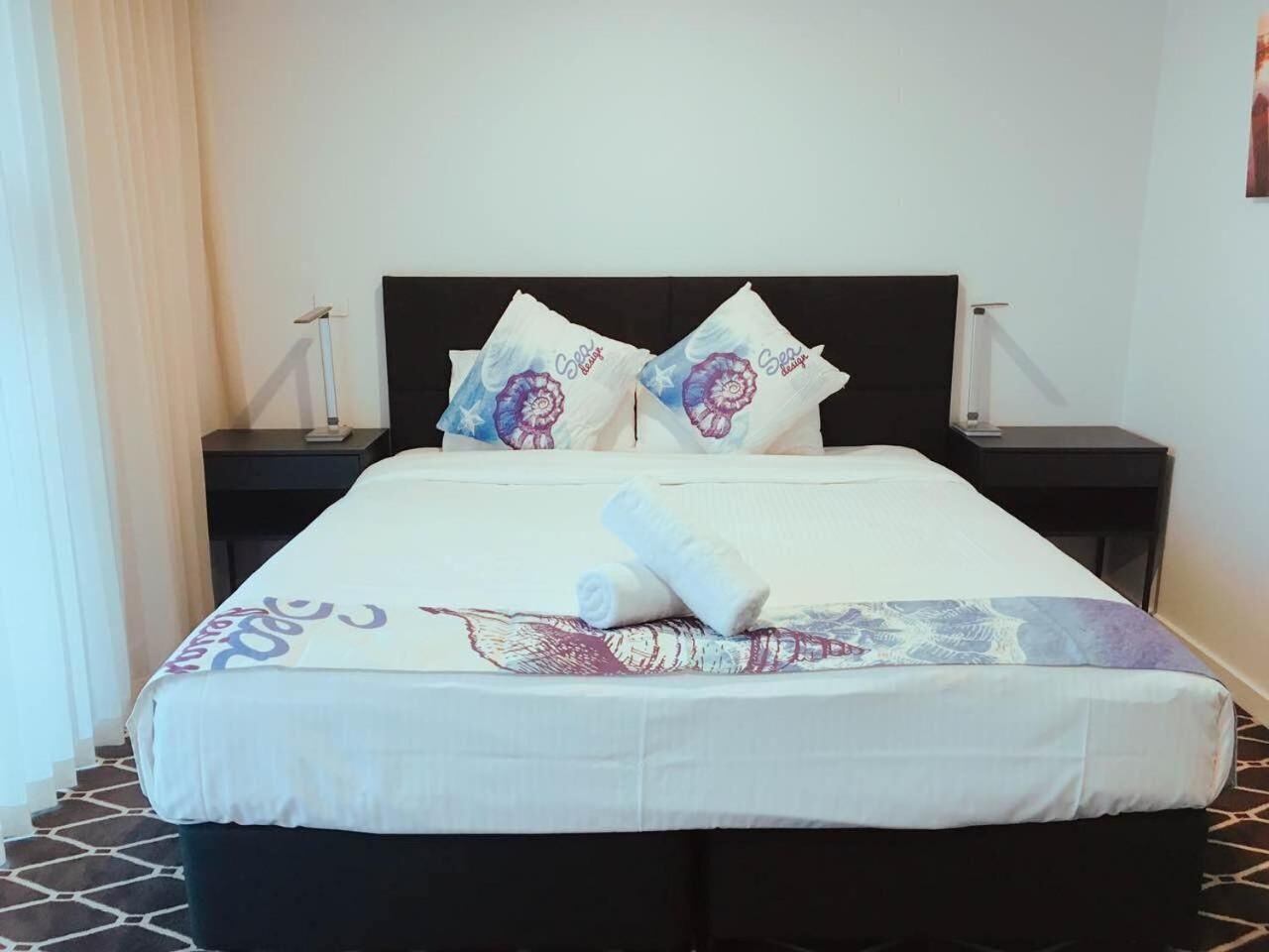 Habitación de hotel con una cama, dos mesitas de noche, una lámpara y un ventanal con cortinas.