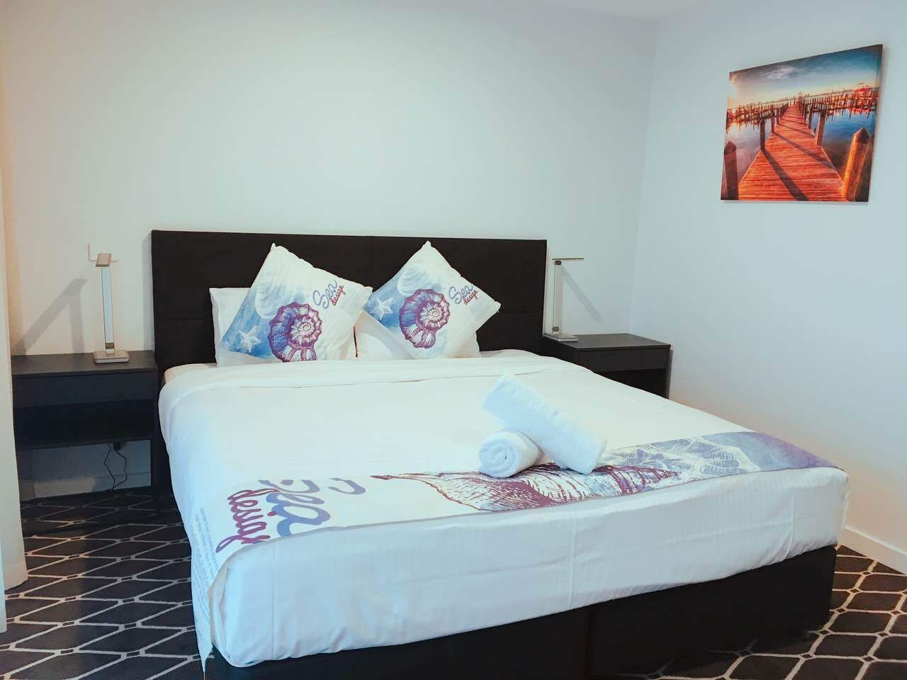 A neatly made bed with a white bedspread and decorative pillows, a bedside table with a lamp, and a painting on the wall.