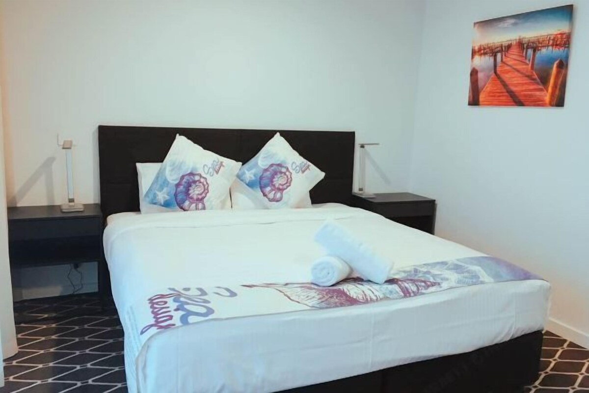 A neatly made bed with a white bedspread and two decorative pillows, a rolled towel on the bed, and a painting of a pier on the wall.