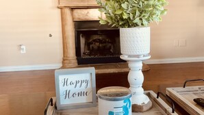 Ein gemütliches Wohnzimmer mit Kamin, Blumentopf, Tablett mit Kerze und einem Schild mit der Aufschrift 'Happy Home'.