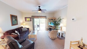 A living room with a brown sofa, a glass coffee table, a ceiling fan, a TV, and a plant.