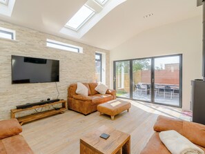 A spacious living room with a large flat-screen TV mounted on a brick wall, a leather sofa, a wooden coffee table, and a sliding glass door leading to an outdoor patio area. - Perfect for creating memories together as a family which will last a lifetime. (Whitby)