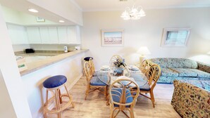 A modern kitchen with white cabinets, a dining area with wicker chairs and a glass table, and a patterned sofa.