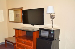 Quarto de hotel com televisão de tela plana na parede, micro-ondas em uma mesinha, cômoda de madeira com gavetas, uma cadeira e um abajur.