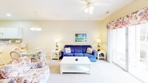 A living room with a glass dining table, floral sofa, blue sofa, white coffee table, and two lamps. There is a ceiling fan, a painting on the wall, and a view through a glass door.