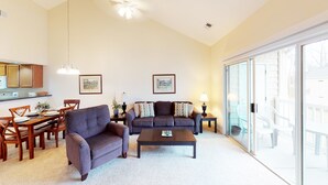A living room with a sofa, armchair, coffee table, and dining area with chairs and a table. There are two framed pictures on the wall, a lamp on a side table, and a sliding glass door leading to an outdoor area.
