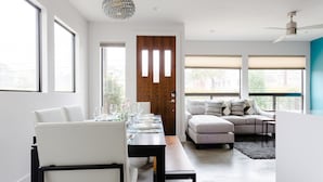 Een moderne woonkamer met een eethoek, met een houten deur, grote ramen, een witte bank en een plafondventilator.