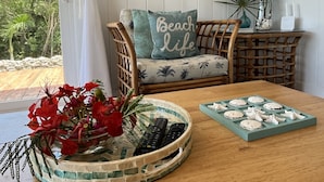 Une chambre décorée sur le thème de la plage, avec un fauteuil en osier, un vase rempli de fleurs rouges, un plateau orné de coquillages et un mur décoré d’œuvres d’art aux motifs tropicaux.