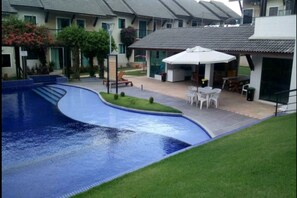 A swimming pool with a curved edge, surrounded by a grassy area and a patio with outdoor furniture. - Condominio Fechado Praia do Atalaia (Salinópolis)
