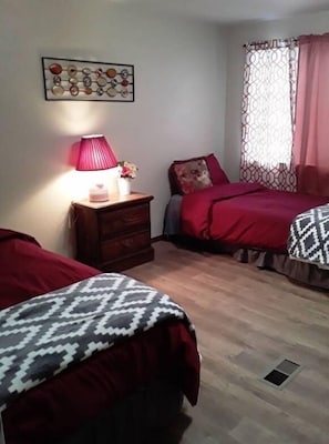 A bedroom with two beds, a bedside table with a lamp, a decorative wall piece, and a window with a curtain. - House  Perfect For Short/Long Term Stays! (Chewelah)