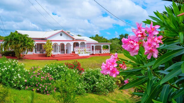 A house with a red and white exterior, a porch, and a garden with various flowers. - Sunny Palm Villas (Laborie)