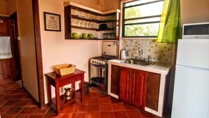 A compact kitchen with a gas stove, sink, and a refrigerator. There is a small table with a basket and a brass pitcher. A framed quote is on the wall, and a window with a green curtain is visible. - Sunny Palm Villas (Laborie)