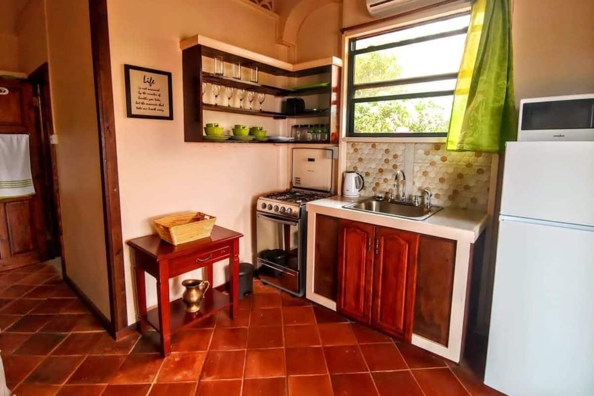 A compact kitchen with a gas stove, sink, and a refrigerator. There is a small table with a basket and a brass pitcher. A framed quote is on the wall, and a window with a green curtain is visible.
