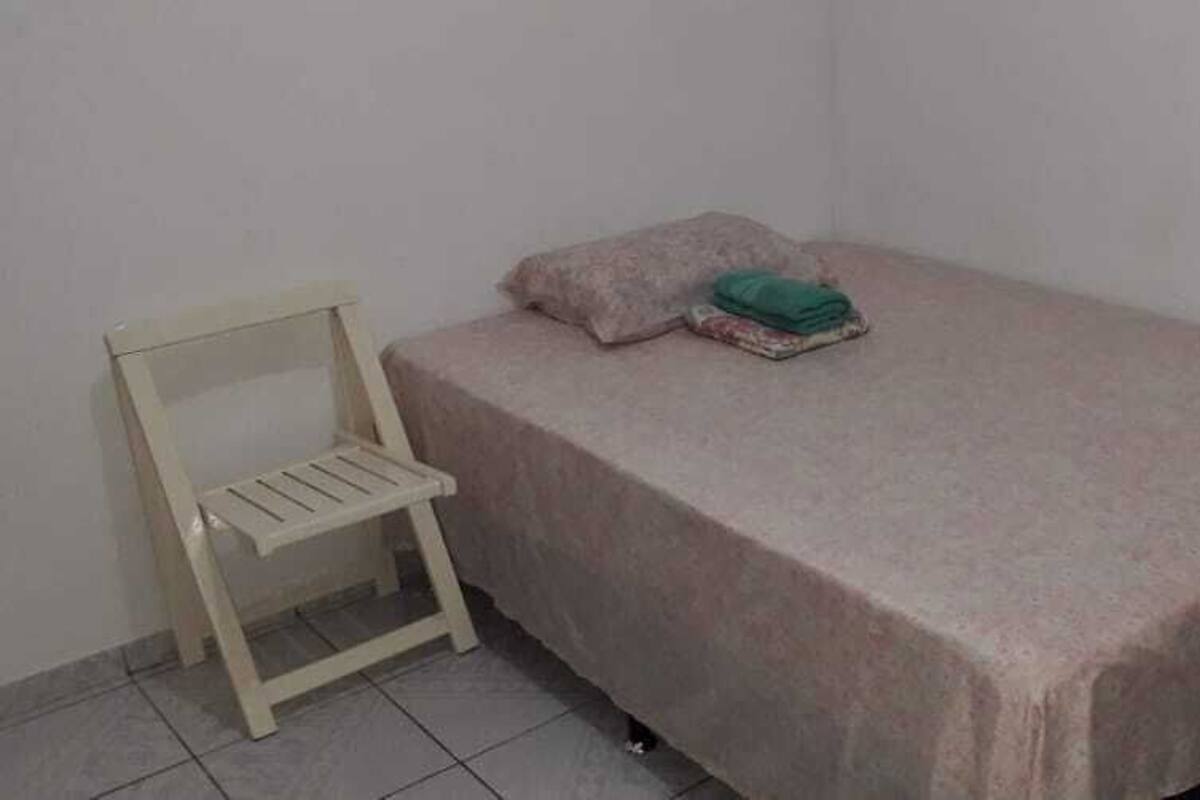A single bed with a pink bedspread, a green folded blanket, and a single pillow on a white wall.
