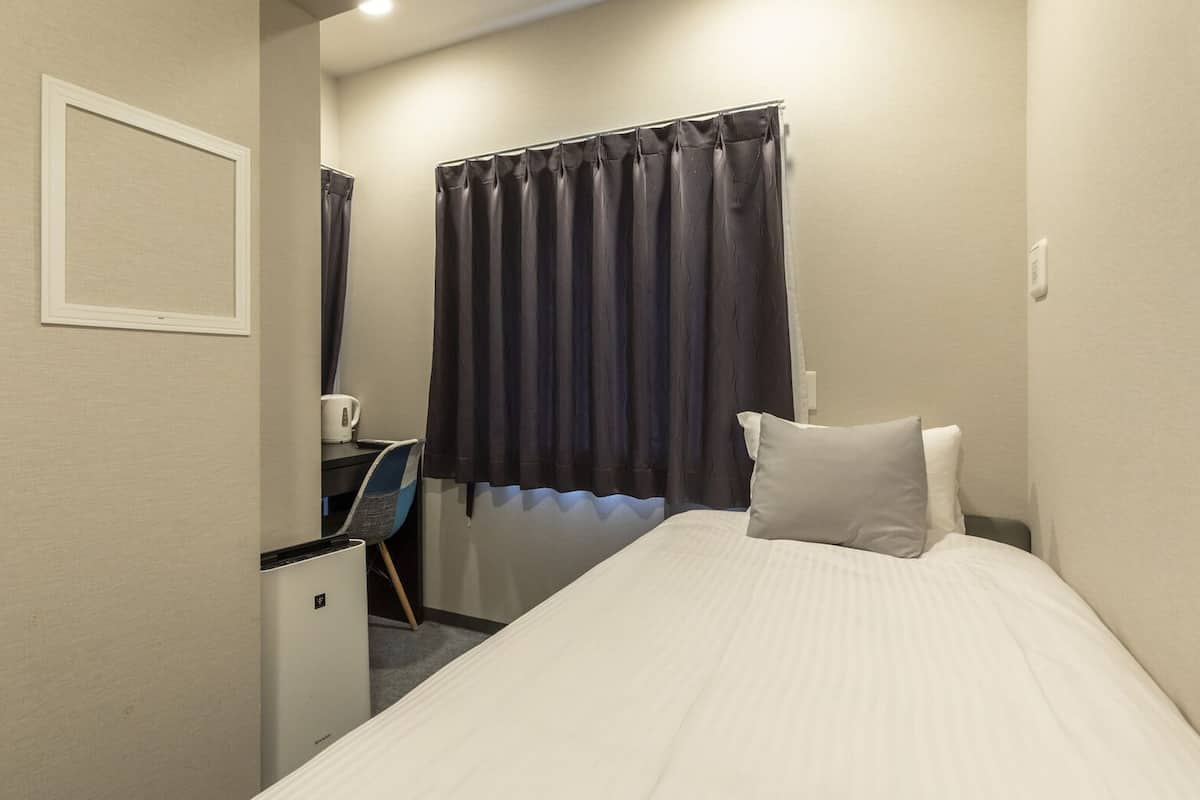 Une petite chambre d’hôtel, bien aménagée et soignée, avec un lit, un bureau et une chaise, une bouilloire et une fenêtre avec des rideaux.