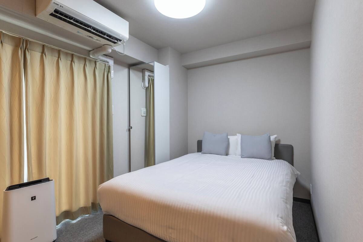 Une chambre d’hôtel comprenant un lit, une table de chevet, un appareil de climatisation et une fenêtre avec des rideaux.
