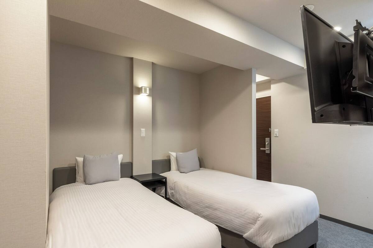 Une chambre d’hôtel avec deux lits simples, une petite table, un téléviseur fixé au mur et une porte donnant sur une salle de bain.