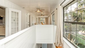Un pasillo largo y estrecho con paredes blancas y suelo de madera, que conduce a una habitación con un ventanal y un ventilador de techo.