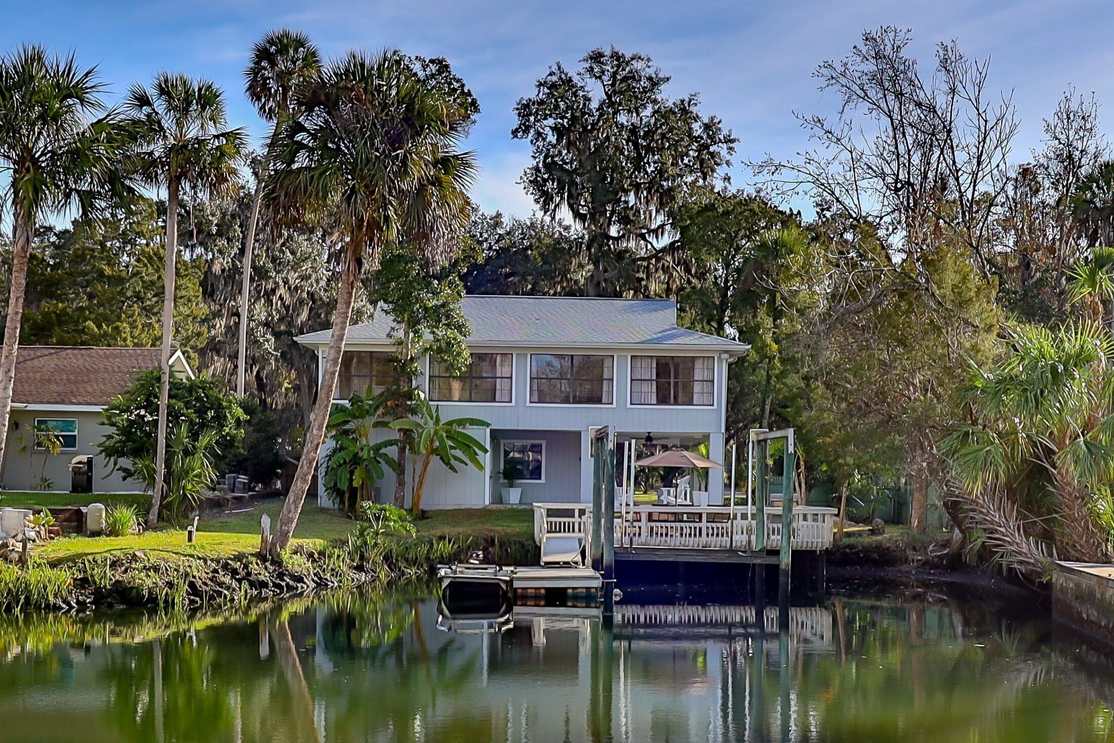 A two-story house with a dock extending into a calm body of water, surrounded by lush greenery and tall trees.